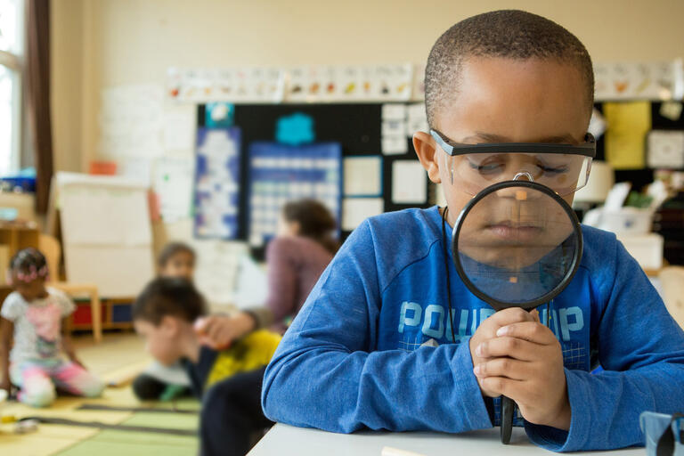A kindergarten student uses a magnifying glass to take a closer look at a pencil.